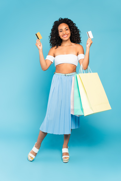 happy african american girl holding credit cards and shopping bags while standing on blue - Foto, Imagem