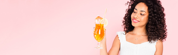 concept panoramique de joyeuse fille afro-américaine regardant le verre avec cocktail isolé sur rose  - Photo, image