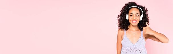 raccolto panoramico di felice ragazza afroamericana in cuffie wireless ascoltare musica e mostrando pollice in su isolato su rosa  - Foto, immagini