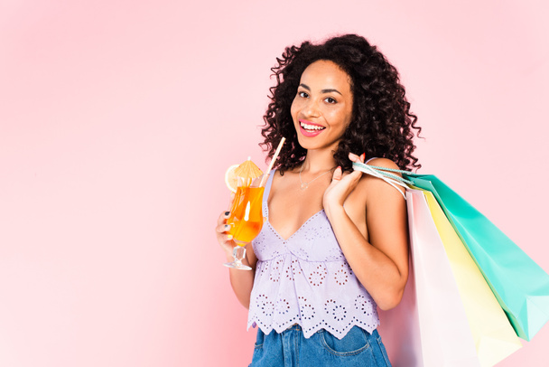 happy african american girl holding shopping bags and cocktail isolated on pink  - Foto, Imagen
