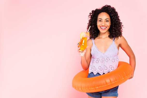 smiling african american woman standing with swim ring and holding cocktail isolated on pink  - Valokuva, kuva