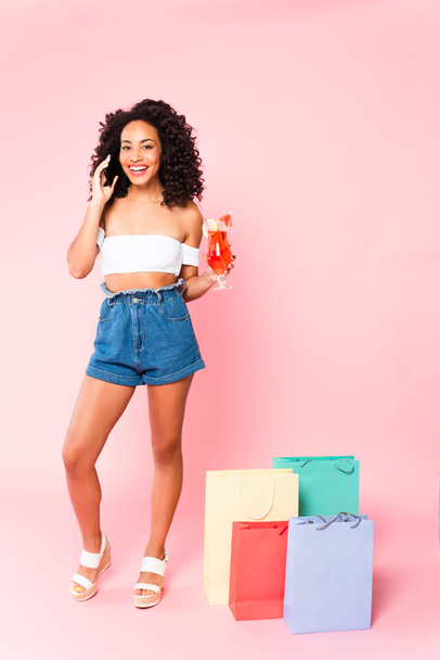 happy african american woman holding cocktail and talking on smartphone while standing near shopping bags on pink  - Foto, Bild