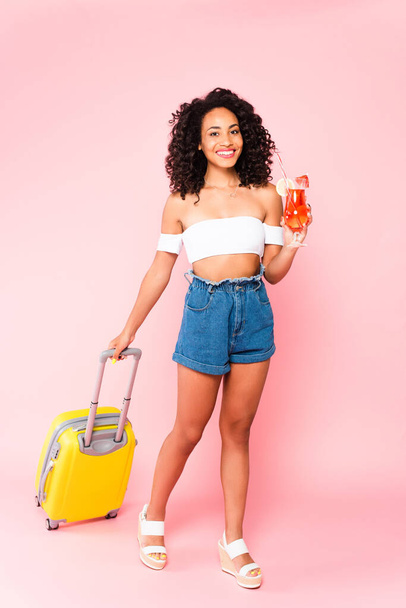 cheerful african american woman holding cocktail and walking with suitcase on pink  - Fotoğraf, Görsel