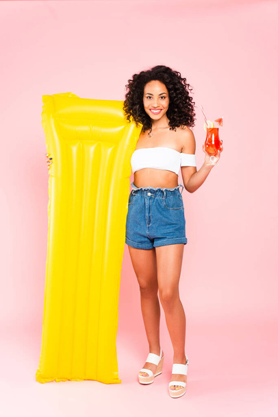 smiling african american girl holding cocktail and inflatable mattress on pink  - Foto, Imagem