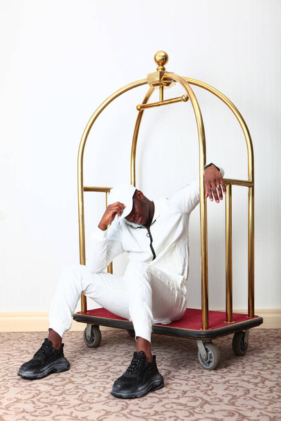 An African-American man in a white suit sits in a Luggage cart. Unrecognizable vertical photo on a white background. The person is resting. Luggage trolley made of yellow metal.Copy space. Fatigue after work. - Foto, Bild