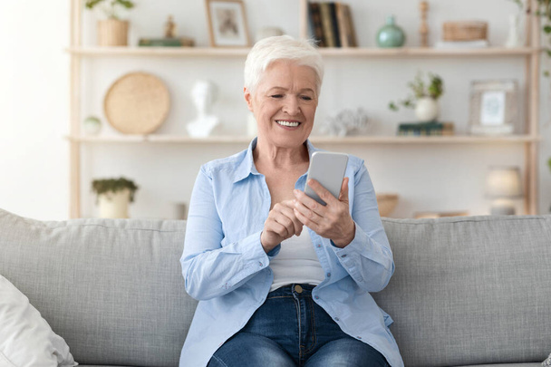 Application de paiement facile. Femme âgée souriante utilisant un téléphone intelligent moderne à la maison - Photo, image