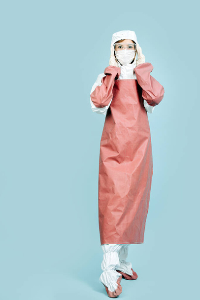 Female surgeon in a white costume covering all skin, wearing red leather apron and sleeves, mask, gloves and glasses. She's fully geared up for surgery. Adjusting glasses. Full length. - Foto, Bild