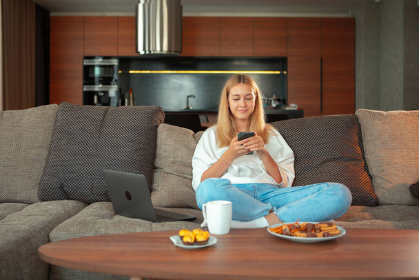 Jolie adolescente souriante assise sur un canapé avec téléphone faisant défiler l'alimentation dans les réseaux sociaux. Environnement familial confortable - Photo, image