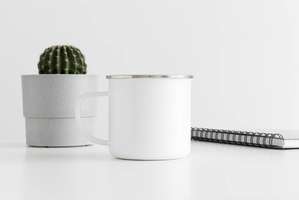 Smalto tazza mockup con un taccuino e un cactus in una pentola sul tavolo bianco. - Foto, immagini