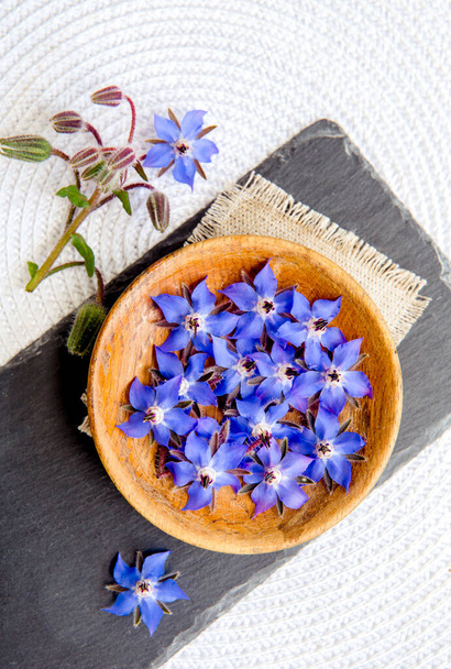 Käyttämällä kaunis tummansininen syötävä kukka elintarvikkeiden koristeluun Borago officinalis (Borago tai borage). Tasainen lay näkymä kukkii puu kulhoon musta kivi levy, valkoinen tausta. - Valokuva, kuva