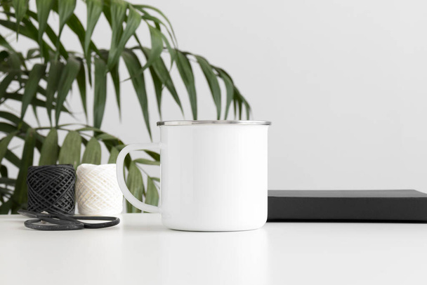 Modèle de tasse en émail avec accessoires d'espace de travail sur une table blanche et une palme. - Photo, image