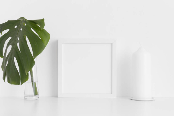 Λευκό τετράγωνο πλαίσιο mockup με ένα φύλλο monstera σε ένα βάζο και ένα κερί σε ένα λευκό τραπέζι. - Φωτογραφία, εικόνα