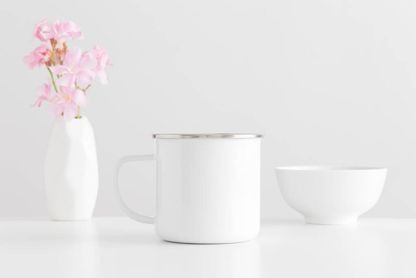 Enamel mug mockup with a bouquet of a pink oleander in a vase and a bowl on a white table. - Fotó, kép