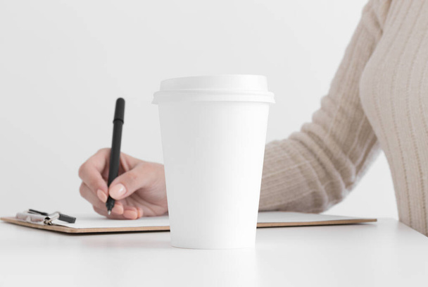 Wit koffie papier beker mockup met vrouw het maken van notities op een klembord. - Foto, afbeelding