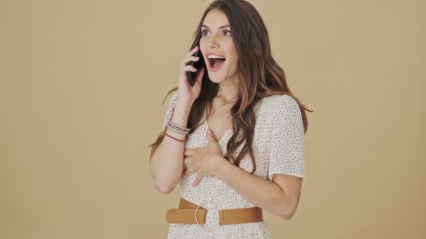 An emotional happy young woman is talking on her smartphone standing over a beige background in studio - Felvétel, videó