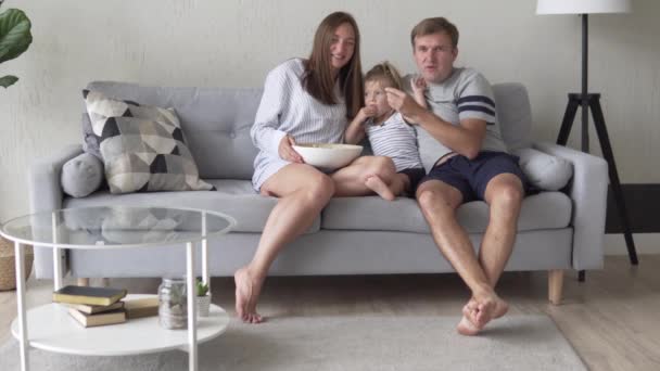 Een gezin met een jong kind kijkt tv en eet popcorn. - Video