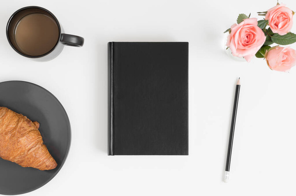 Top kilátás egy fekete könyv mockup egy csokor rózsaszín rózsa, kávé és croissant egy fehér asztalon. - Fotó, kép
