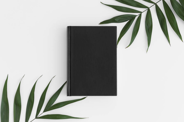 Top view of a black book mockup with palm leaf decoration on a white table. - Photo, Image