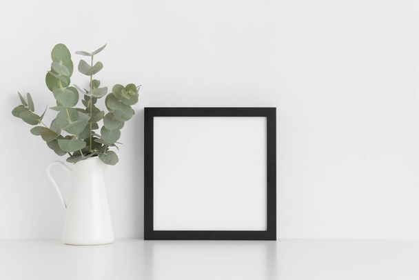 Schwarze quadratische Rahmen-Attrappe mit einem Strauß Eukalyptus in einer Vase auf einem weißen Tisch.. - Foto, Bild