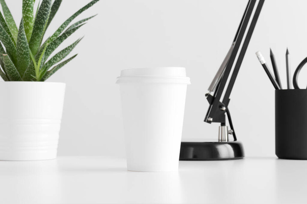 Coffee paper cup mockup with a cactus in a pot, lamp and workspace accessories on a white table. - Valokuva, kuva