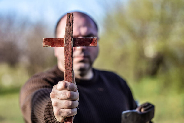 Ένας άντρας κρατά ένα ξύλινο χριστιανικό σταυρό μπροστά του, ένα τσεκούρι στο άλλο του χέρι. Η έννοια της εκρίζωσης δαιμόνων στις Απόκριες. - Φωτογραφία, εικόνα