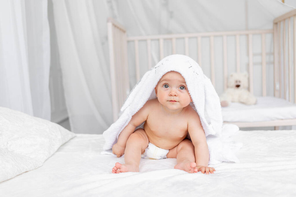 Egy 6 hónapos kislány egy fehér ágyon ül egy törülközőben egy pelenkás gyerekszobában fürdés után. - Fotó, kép