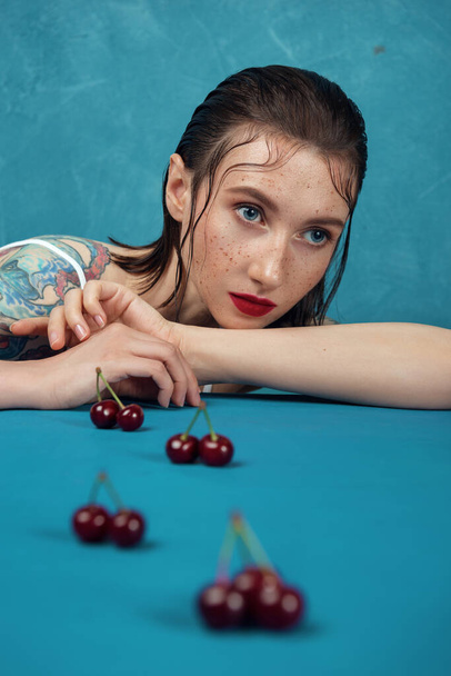 Молодая модель, сидящая за столом с вишневыми ягодами на синем фоне.  - Фото, изображение