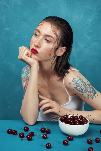 Jeune femme tatouée en chemise blanche assise à la table avec des baies de cerises devant le fond bleu.  - Photo, image
