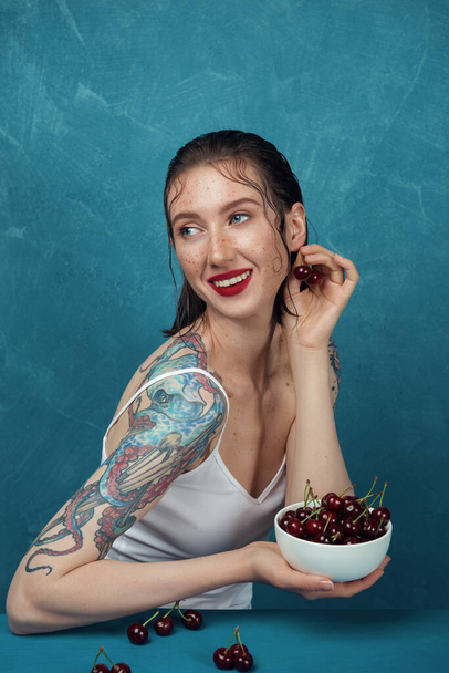 Young happy freckled woman sits at the table with cherry bowl in front of the blue background.  - 写真・画像