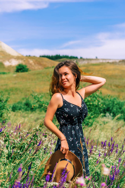 A happy young woman in a dress holds a hat against a background of blooming wildflowers and tall grass in a hilly area - Photo, Image