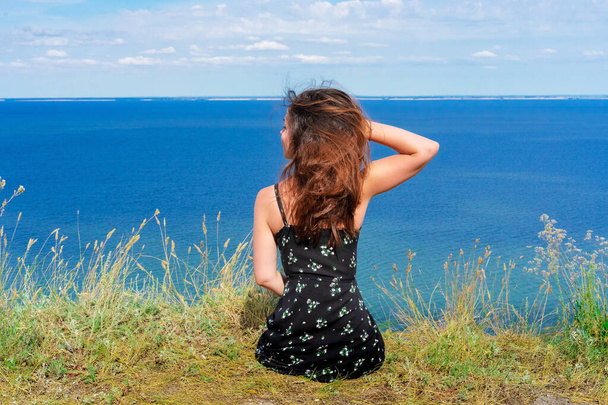  Une jeune femme heureuse dans une robe est assise sur une falaise, dos à la caméra, avec la mer bleue devant elle - Photo, image