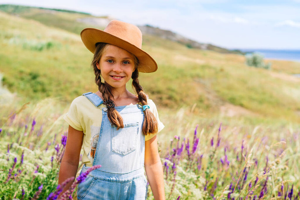 Portrait of a charming little girl in her mother's hat holding apples in her hand against a picturesque hilly landscape with a lake - Foto, Imagem