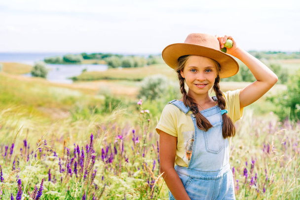 Portrait of a charming little girl in her mother's hat holding apples in her hand against a picturesque hilly landscape with a lake - Foto, Imagem