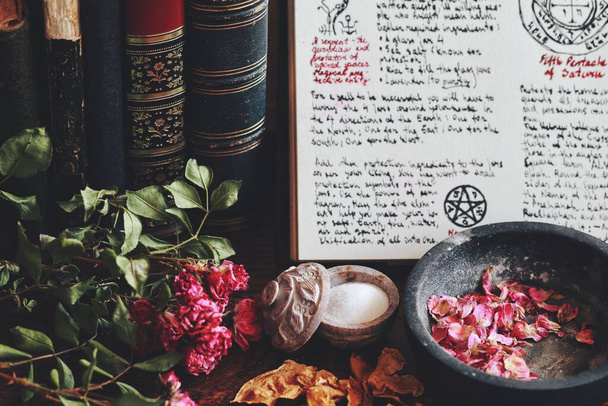 Wiccan witch altar with an open book of shadows with hand written spell in it, ready for spell casting. Grimoire page with symbols and drawings. Old vintage books, dried flowers, salt ingredients in the background - Photo, Image