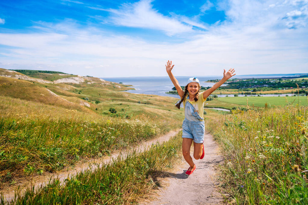Ένα χαρούμενο κοριτσάκι, ένα παιδί με καπέλο και τζιν φόρμα τρέχει στην κάμερα σε μια γραφική περιοχή με λόφους. - Φωτογραφία, εικόνα