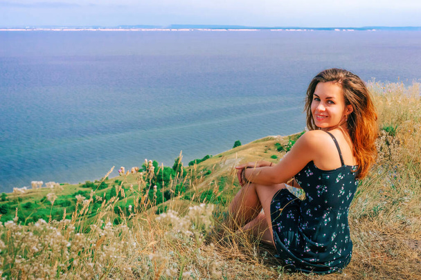 Une jeune femme heureuse dans une robe est assise sur une falaise, dos à la caméra, avec la mer bleue devant elle - Photo, image