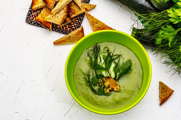 Green zucchini cream soup. Healthy vegan (vegetarian) food, crispy toast, sesame seeds, whole ripe fruits, fresh greens. White putty background, top view - Foto, imagen