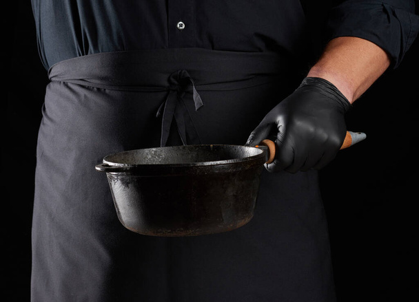 cook in black uniform and latex gloves holds an empty round vintage black cast iron pan in front of him, low key - Photo, image