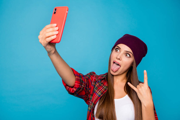 Closeup foto docela bláznivá dáma držet telefon blogger dělat selfies ukazující rohy ulice vzhled nový portrét nosit příležitostný klobouk kostkovaný košile izolované modré barvy pozadí - Fotografie, Obrázek