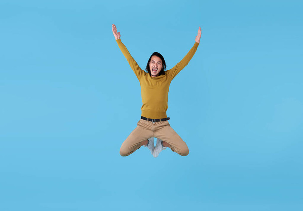 Happy energetic young Asian man jumping in mid-air isolated on blue background. - Фото, изображение