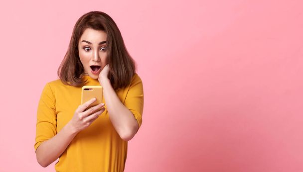 счастливая и удивительная молодая женщина хорошо выглядит, используя мобильный телефон на розовом фоне пространства копирования. - Фото, изображение