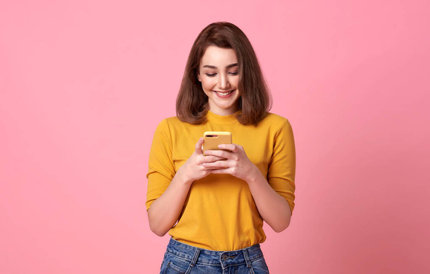щаслива молода жінка добре виглядає, використовуючи мобільний телефон ізольовано на рожевому фоні
. - Фото, зображення