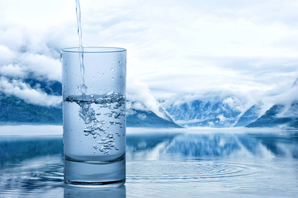 Ρίχνει νερό σε ένα ποτήρι ενάντια στο φυσικό τοπίο με τη λίμνη και τα βουνά - Φωτογραφία, εικόνα