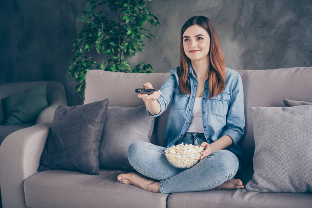 Portrait of positive pretty girl sit divan legs crossed watch interesting movie switch channels eat popcorn barefoot in house indoors - Фото, изображение