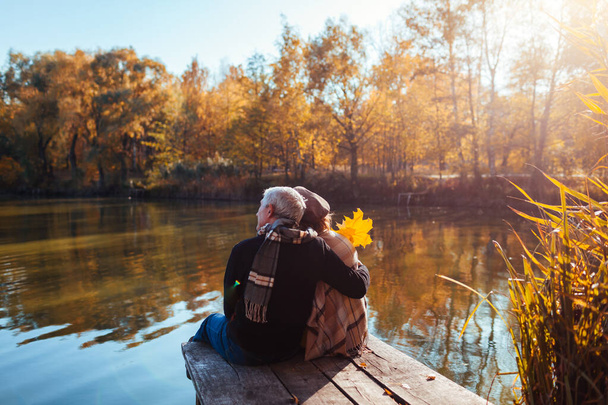 Senior family couple relaxing by autumn lake. Man and woman enjoying natural landscape and hugging sitting on pier - Foto, immagini
