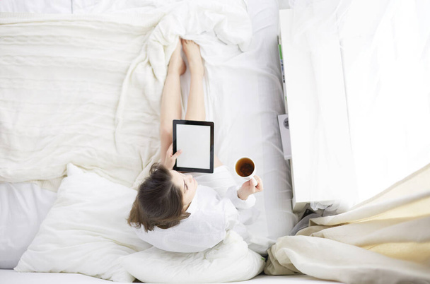 Portrait of woman using tablet and drinking coffee while sitting on the bed in the morning - Valokuva, kuva