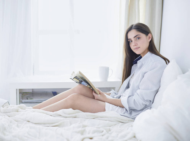 Portrait of woman using tablet and drinking coffee while sitting on the bed in the morning - 写真・画像