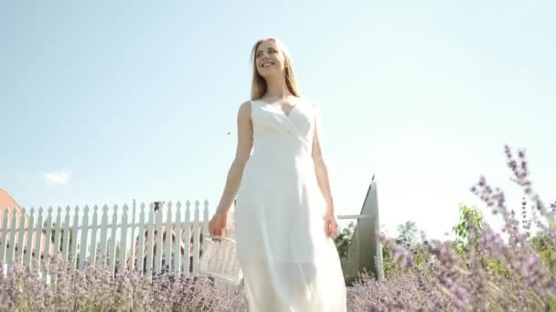 Caucasian 25-year-old beautiful blonde in a long white dress walks on a lavender field on a sunny summer evening with a basket in her hands. Many purple flowers. Back shot. Lavender - Felvétel, videó