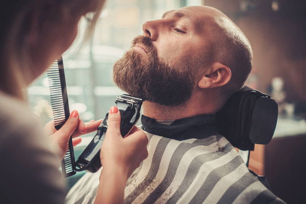 bel homme sérieux assis sur la chaise tandis que le styliste coupe sa barbe. - Photo, image