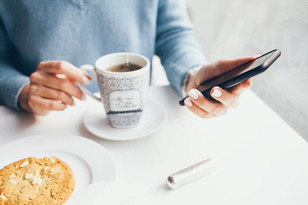 Primer plano de las manos de mujer con manicura blanca sosteniendo el teléfono móvil y desayunando al aire libre en un agradable café. Leer noticias, redes sociales, navegar por la web / Internet en el teléfono inteligente. Enfoque selectivo
 - Foto, imagen